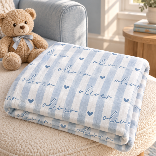 A folded baby blanket with light blue and white vertical stripes, featuring the name 'oliver' in script and small hearts, displayed on a beige armchair next to a teddy bear in a softly lit nursery setting.