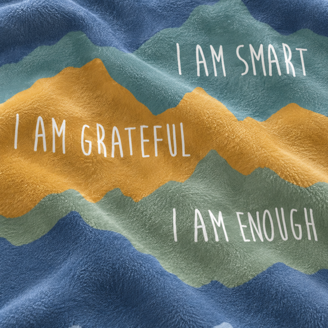 A close-up of a textured fleece blanket with a mountain pattern in shades of blue, teal, and mustard yellow, featuring white text that reads 'I AM SMART', 'I AM GRATEFUL', and 'I AM ENOUGH'.