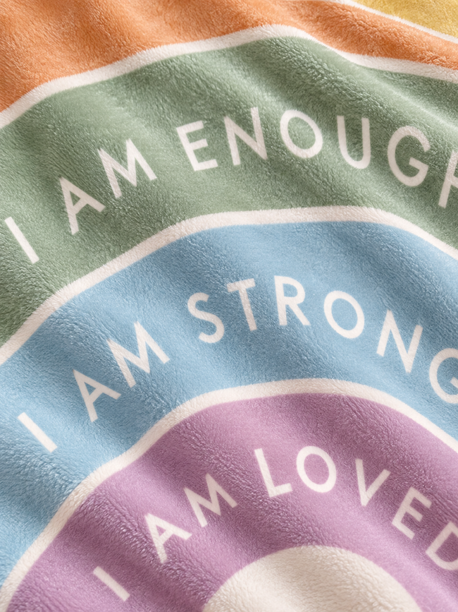 Close-up of a plush fleece blanket with a rainbow design and the words 'I AM ENOUGH', 'I AM STRONG', and 'I AM LOVED' in white text.