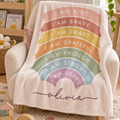 A personalized fleece throw blanket draped over a cream armchair in a cozy child's room. The blanket is cream-colored with a rainbow graphic made of pastel stripes and arches containing positive affirmations like 'I AM KIND', 'I AM BRAVE', 'I AM GRATEFUL', 'I AM ENOUGH', 'I AM STRONG', and 'I AM LOVED', finished with the name 'Olivia' in a dark purple script at the bottom.