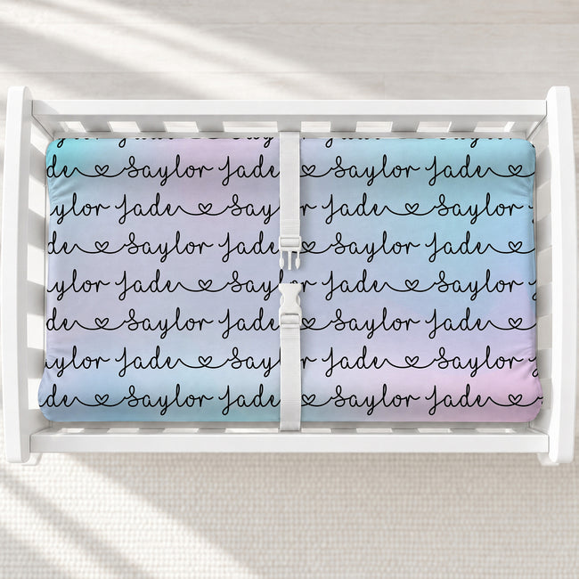 A personalized changing pad cover featuring a pastel blue, purple, and pink gradient background with the names 'Saylor Jade' repeated in black script with small heart motifs.