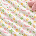 A close-up of a plush white blanket with a repeating pattern of pastel pink, green, and yellow stars, featuring the name 'Anna' printed in pink script. A soft pink plush star toy is partially visible in the top right corner.