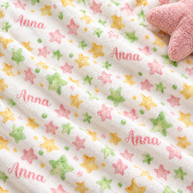 A close-up of a plush white blanket with a repeating pattern of pastel pink, green, and yellow stars, featuring the name 'Anna' printed in pink script. A soft pink plush star toy is partially visible in the top right corner.