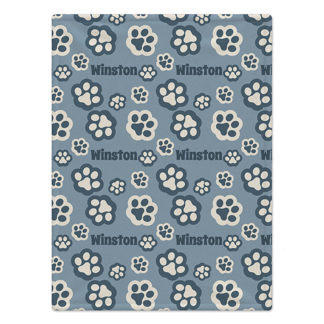 A personalized dog blanket featuring a  repeating pattern of cream-colored paw prints and the word 'Winston' in dark gray on a dusty blue background.
