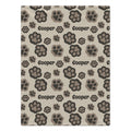 A personalized dog blanket featuring a  repeating pattern of dog paw prints in dark brown, gray, and beige on a light beige background, with the name 'Cooper' printed in black text.