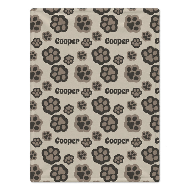 A personalized dog blanket featuring a  repeating pattern of dog paw prints in dark brown, gray, and beige on a light beige background, with the name 'Cooper' printed in black text.