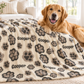 A golden retriever dog lies on a cream pet blanket with gray and taupe paw prints and the name 'Cooper' printed repeatedly, in a bright living room setting.