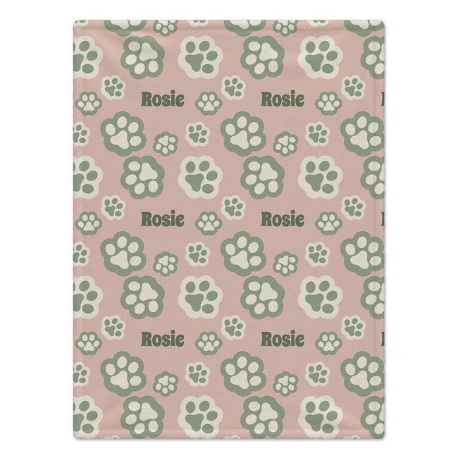 A personalized dog blanket featuring a repeating pattern of green paw prints and the name 'Rosie' on a dusty pink background.