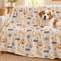A dog lying on a couch covered with a cream pet blanket printed with bones, paw prints, bowls, hearts, and the word "SPOT" repeated in dark blue. The dog is a white and tan mixed breed.