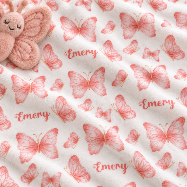 White fabric with a repeating pattern of pink butterflies and the word 'Emery' in pink script, shown close up with a plush pink butterfly toy partially visible in the top left corner.