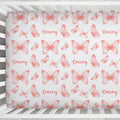 A crib mattress or fitted sheet printed with a repeating pattern of coral-pink butterflies and the name 'Emery' in script, shown inside a white wooden crib.