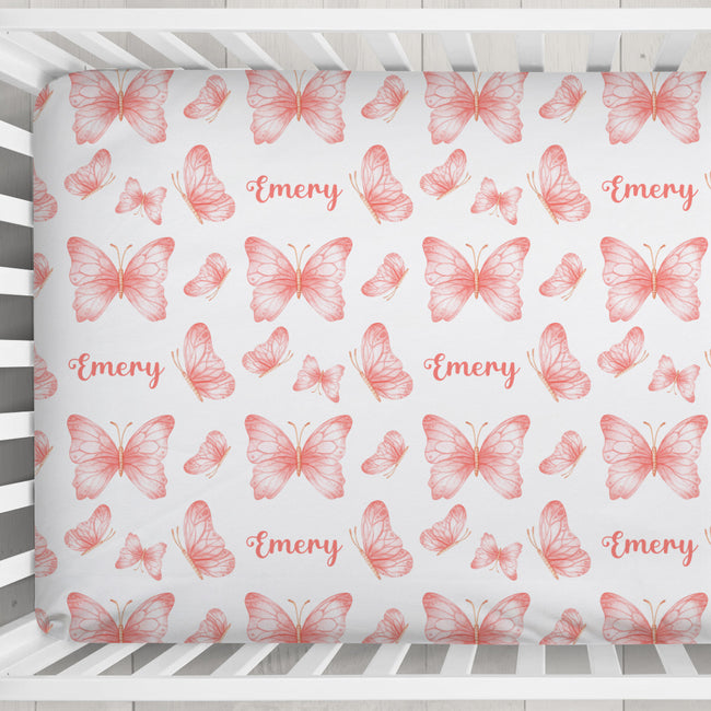 A crib mattress or fitted sheet printed with a repeating pattern of coral-pink butterflies and the name 'Emery' in script, shown inside a white wooden crib.