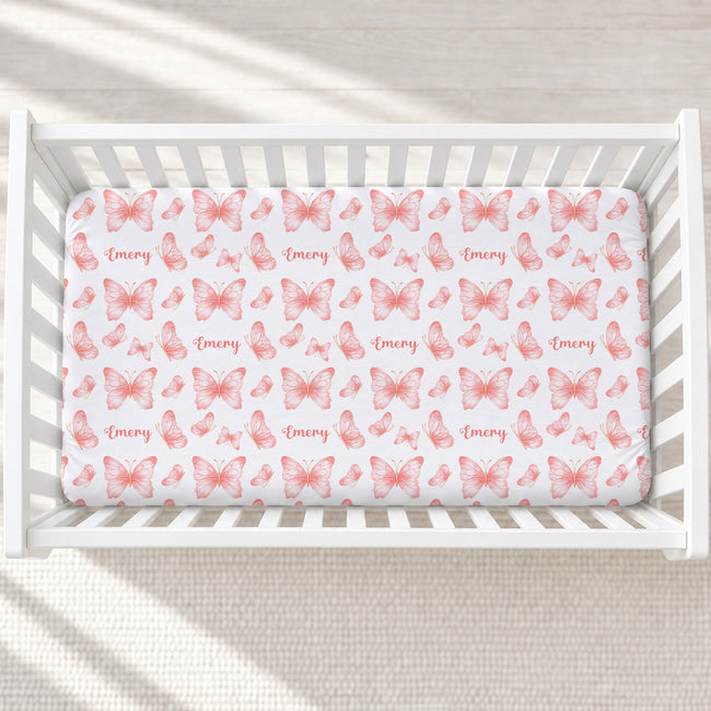 A white crib mattress fitted sheet with a repeating pattern of pink butterflies and the name 'Emery' in script, shown inside a white wooden crib on a light textured rug.