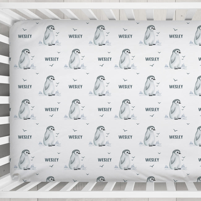 A white crib mattress fitted sheet featuring a repeating pattern of penguin illustrations, small birds, and the name 'WESLEY' printed in dark gray.