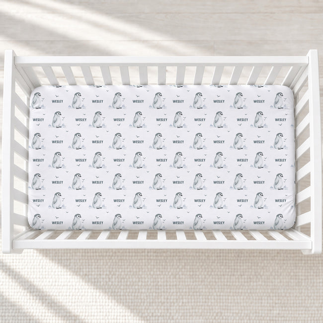 A white crib mattress fitted sheet with a repeating pattern of penguin illustrations and the name 'WESLEY' printed in black, shown inside a white wooden crib.