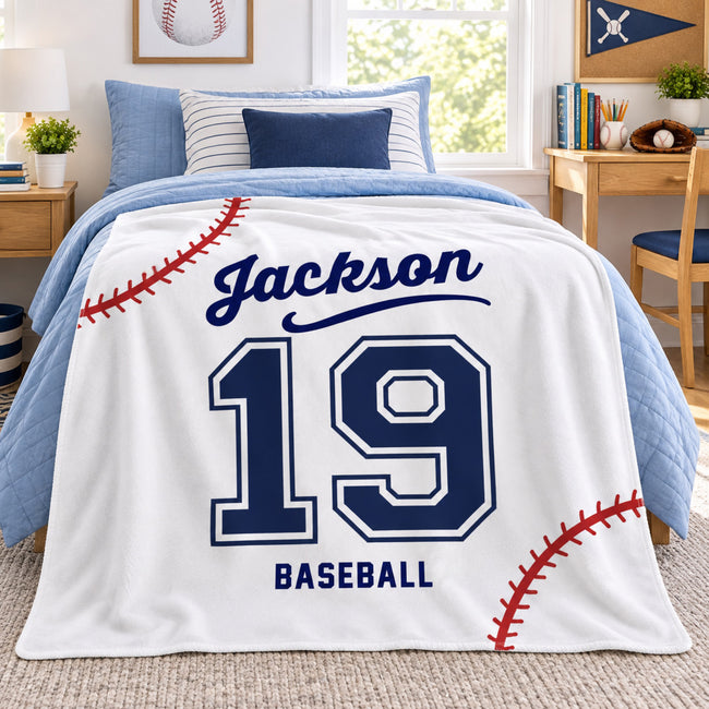 A personalized baseball-themed blanket draped over a bed in a bright, modern bedroom. The blanket is white with navy blue text reading 'Jackson' and 'BASEBALL', the number '19' in navy and white, and red baseball-stitch graphics.