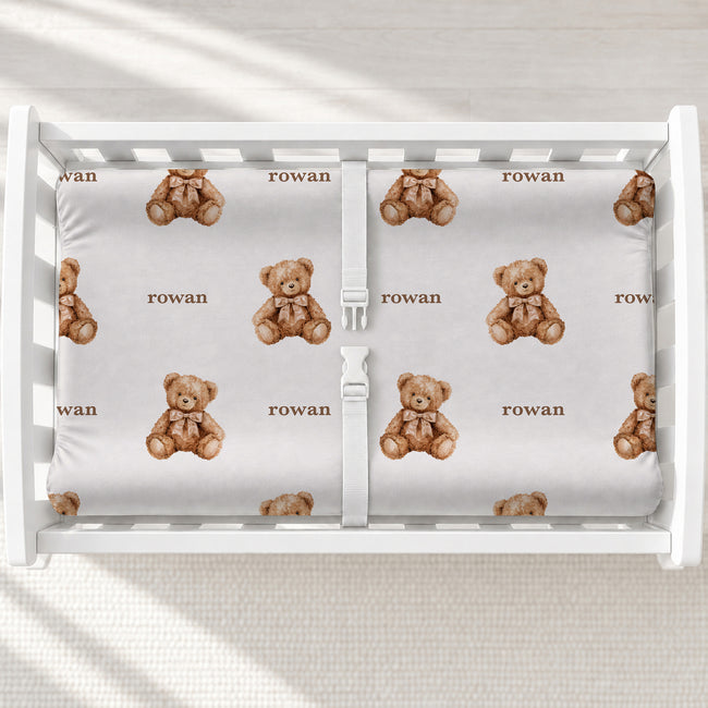 Personalized teddy bear changing pad cover with watercolor bears wearing bows and the name Rowan in brown, shown on a white changing table.