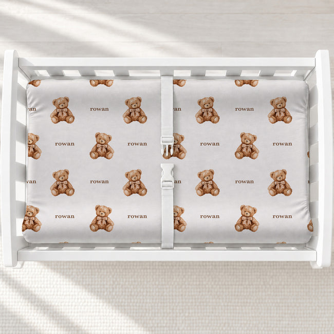 A top-down view of a white baby changing table containing a changing pad cover with a repeating pattern of small brown teddy bears and the word 'Rowan' printed underneath each bear.