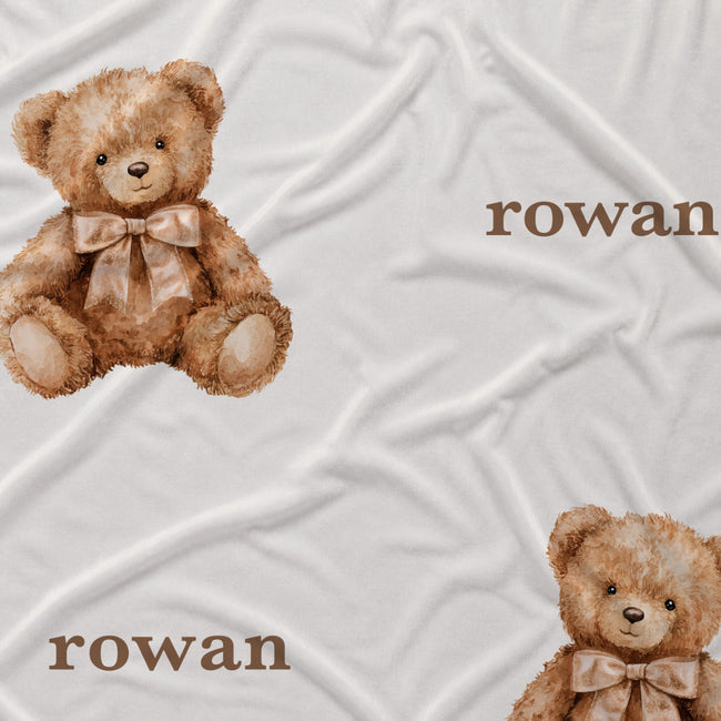 Close-up of watercolor teddy bears wearing beige bows beside the name Rowan in brown lettering on white fabric.