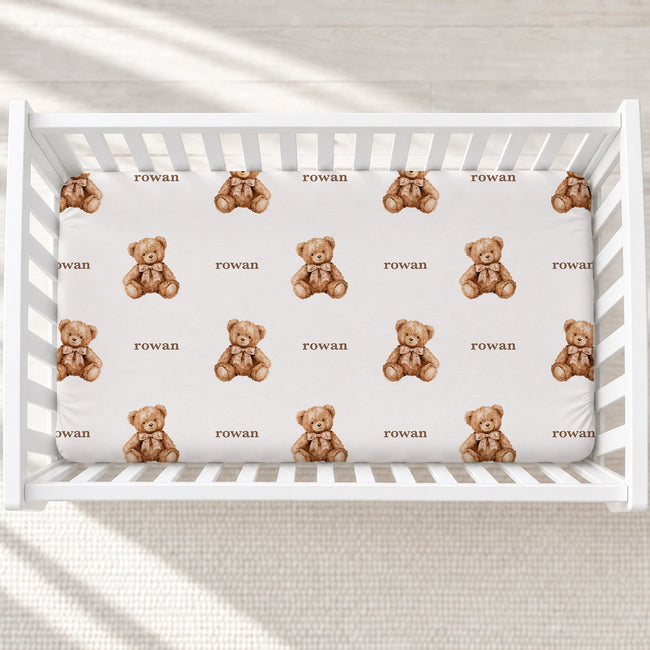 Personalized teddy bear crib sheet with brown watercolor bears, beige bows, and the name Rowan, shown in a white crib.