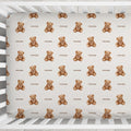 A top-down view of a white wooden baby crib containing a fitted sheet with a repeating pattern of small brown teddy bears and the word 'Rowan' printed underneath each bear.