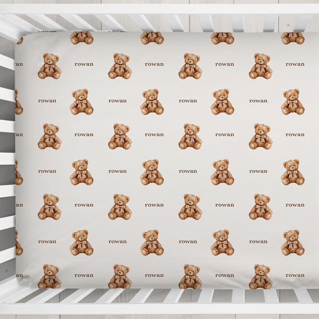 A top-down view of a white wooden baby crib containing a fitted sheet with a repeating pattern of small brown teddy bears and the word 'Rowan' printed underneath each bear.