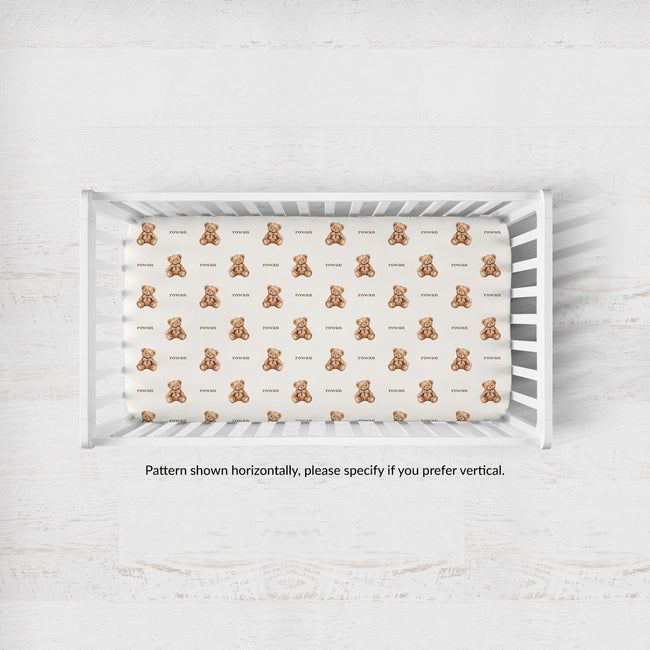 A top-down view of a white wooden baby crib containing a fitted sheet with a repeating pattern of small brown teddy bears and the word 'Rowan' printed underneath each bear.