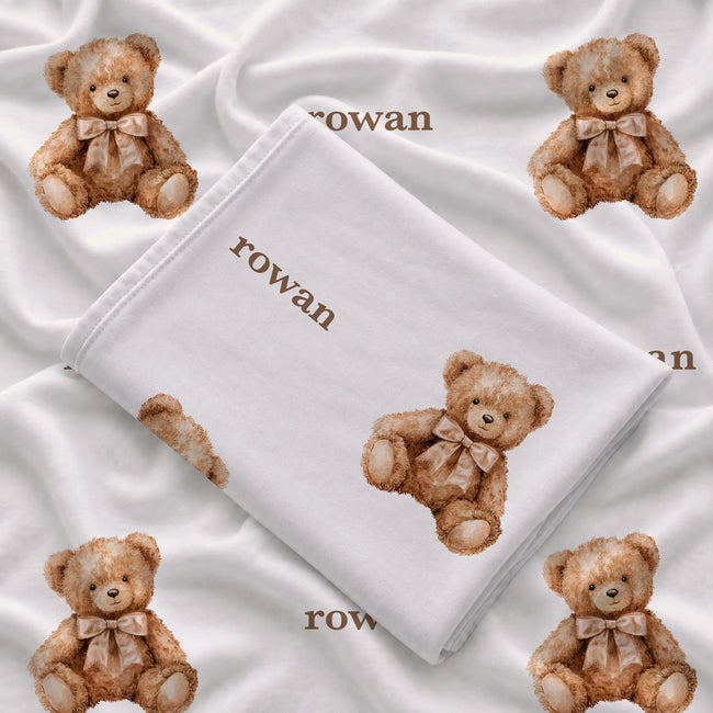 Folded teddy bear swaddle blanket showing the name Rowan in brown and watercolor bears wearing beige bows on white fabric.