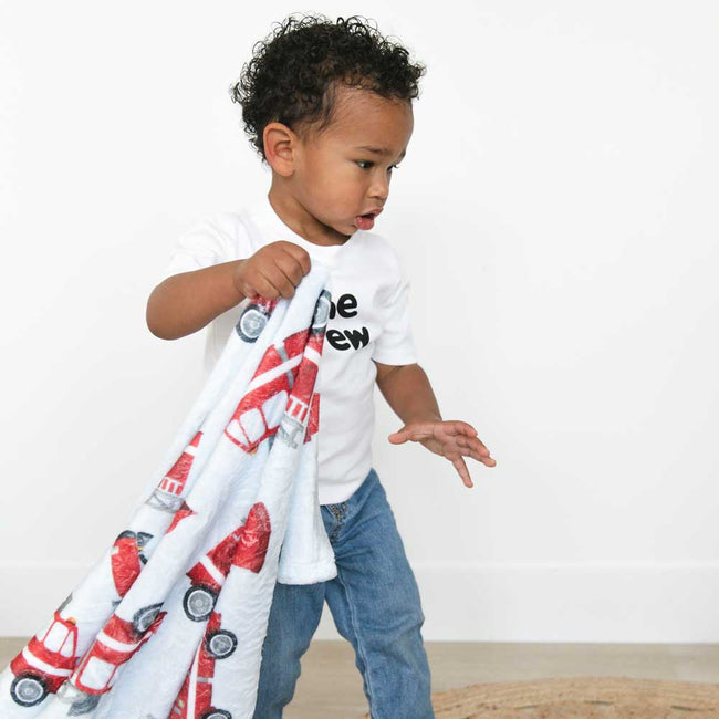 A toddler wearing a white graphic t-shirt and blue jeans holds a baby blue personalized blanket with a red fire truck pattern against a plain white wall.