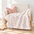 A personalized blanket draped over a cream armchair in a bright nursery setting. The blanket has a white background with a colorful watercolor floral pattern in pink, yellow, and blue, featuring the names 'AMELIA' and 'Grace' printed in script.