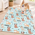 A corgi dog stands on a light blue pet blanket printed with cartoon dogs, paw prints, bones, and the name 'Lacey' in a bright room with wood flooring and a sofa nearby.