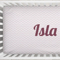 A fitted crib sheet with a light pink and white chevron pattern and the name 'Isla' printed in dark mauve script, shown inside a white wooden crib.