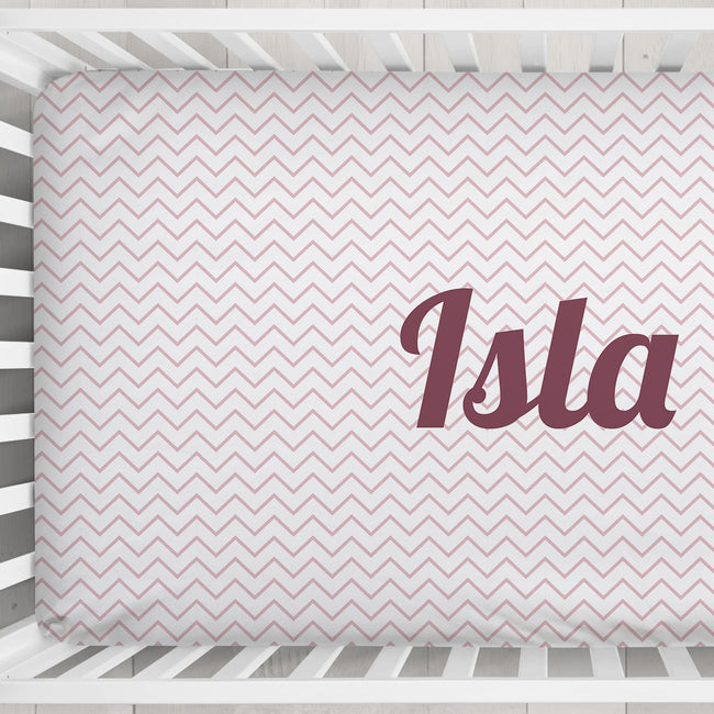 A fitted crib sheet with a light pink and white chevron pattern and the name 'Isla' printed in dark mauve script, shown inside a white wooden crib.