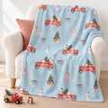 A light blue fleece throw blanket with a Christmas-themed print of red trucks, snowmen, and trees, draped over a cream armchair with a pink pillow.
