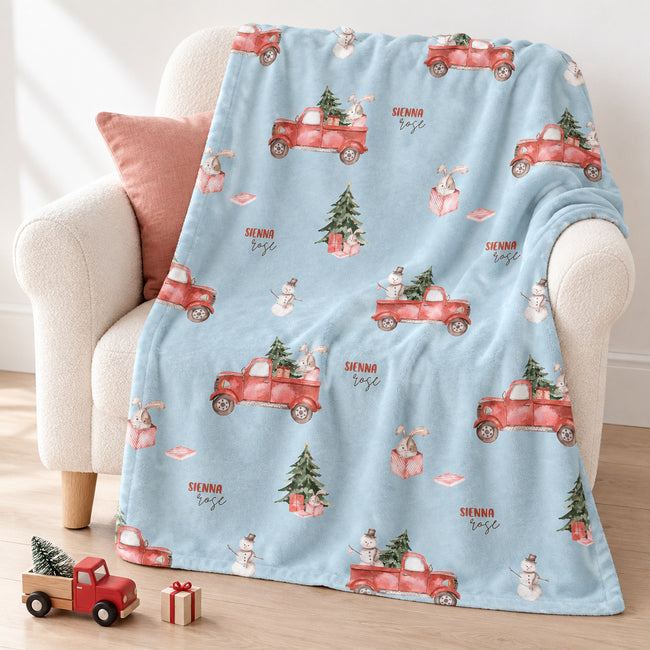 A light blue fleece throw blanket with a Christmas-themed print of red trucks, snowmen, and trees, draped over a cream armchair with a pink pillow.