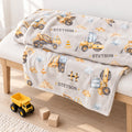 A personalized blanket with a construction theme, featuring the name 'STETSON' and yellow construction vehicles, draped over a light wood bench in a bright room with a toy truck and blocks on the floor.