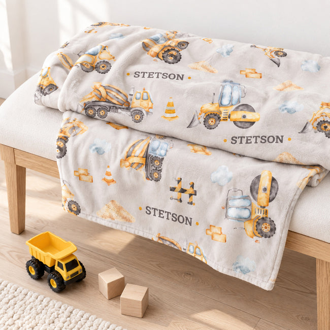A personalized blanket with a construction theme, featuring the name 'STETSON' and yellow construction vehicles, draped over a light wood bench in a bright room with a toy truck and blocks on the floor.