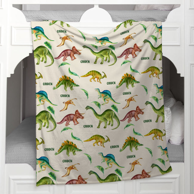 A fleece throw blanket with a colorful dinosaur pattern is draped over the headboard of a white bed.