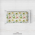 A top-down view of a white wooden crib on a white wood floor, featuring a colorful dinosaur-patterned mattress protector or fitted sheet. The sheet has a cream background with various dinosaur illustrations in green, yellow, blue, and red, each labeled with its species name.