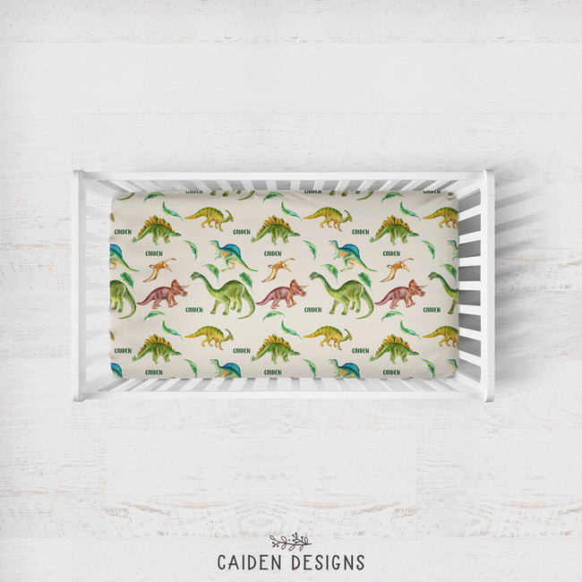 A top-down view of a white wooden crib on a white wood floor, featuring a colorful dinosaur-patterned mattress protector or fitted sheet. The sheet has a cream background with various dinosaur illustrations in green, yellow, blue, and red, each labeled with its species name.