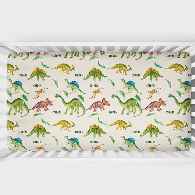A crib mattress fitted sheet with a colorful dinosaur pattern on a cream background, shown inside a white wooden crib.