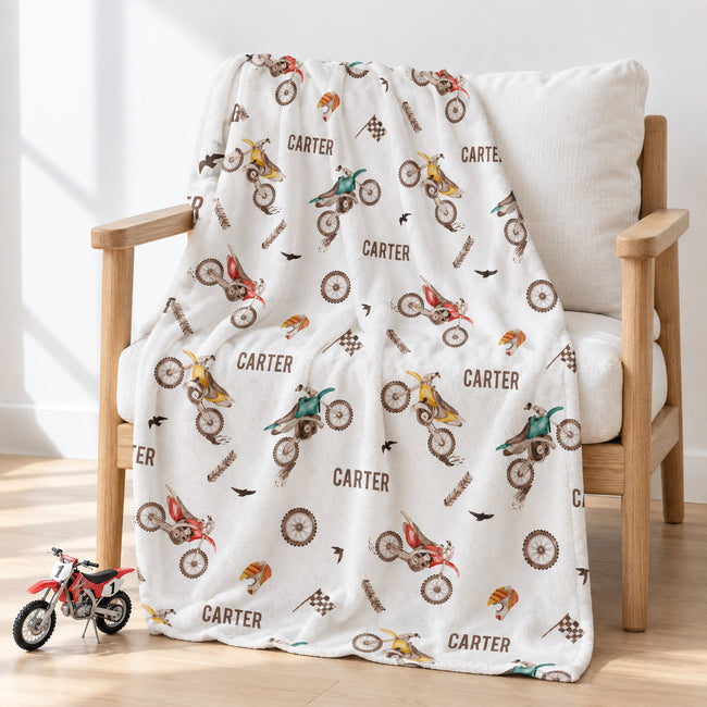 A cream-colored throw blanket with a colorful dirt bike and motorcycle pattern and the name 'CARTER' printed repeatedly, draped over a light wood armchair in a bright room.