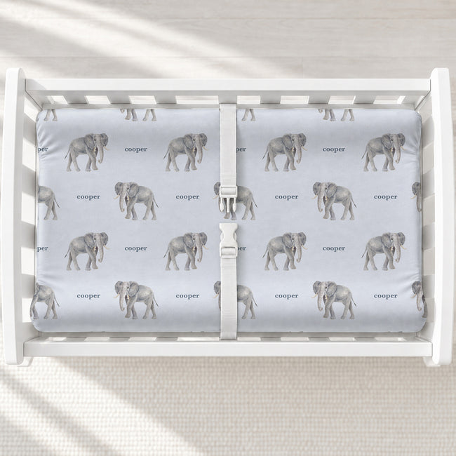 Little Elephant changing pad cover with gray watercolor elephants and the name Cooper on pale blue, shown on a white changing table.