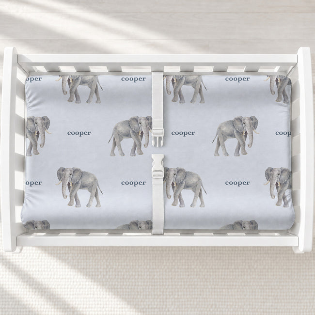 Little Elephant changing pad cover with gray watercolor elephants and the name Cooper on pale blue, shown on a white changing table.