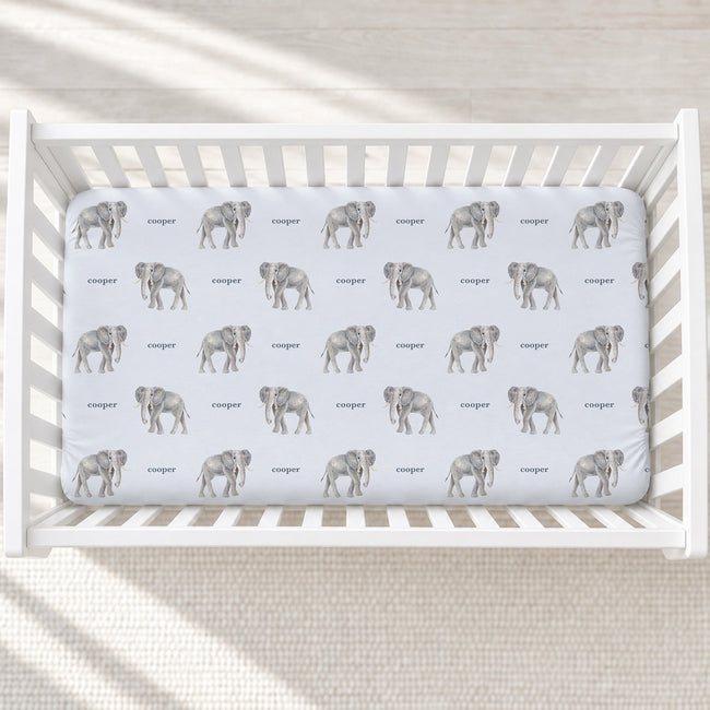 A white wooden crib with a fitted sheet featuring a repeating pattern of gray elephants and the word 'Cooper' in gray text, photographed from above on a light textured floor.