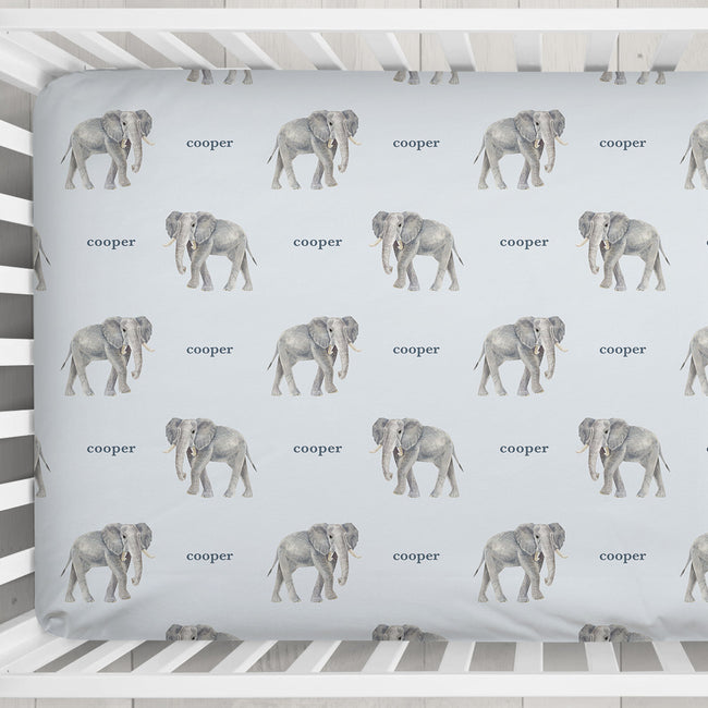 A white wooden crib with a fitted sheet featuring a repeating pattern of gray elephants and the word 'Cooper' in gray text, photographed from above on a light textured floor.