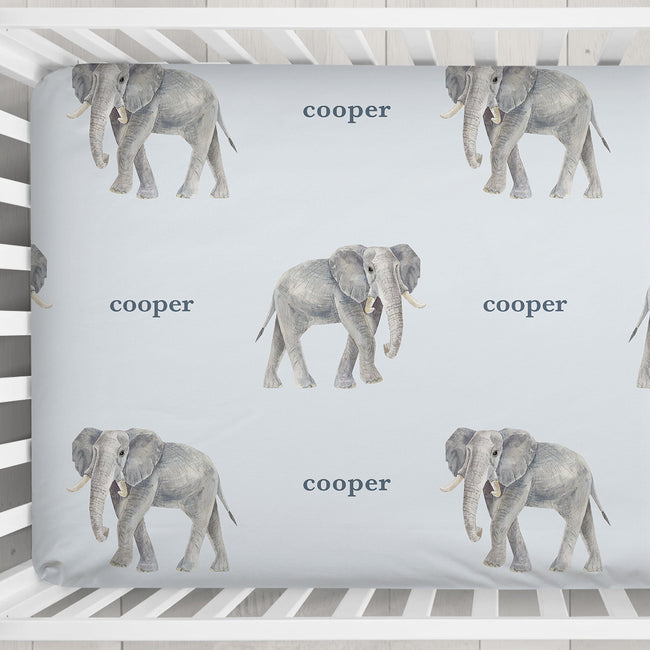 Close-up of Little Elephant crib sheet showing gray watercolor elephants and Cooper in slate blue lettering on a pale blue background.