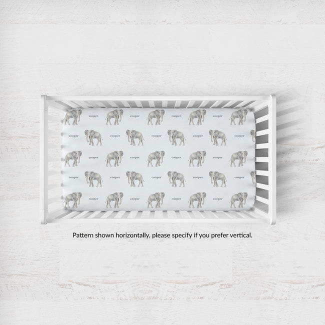 A white wooden crib with a fitted sheet featuring a repeating pattern of gray elephants and the word 'Cooper' in gray text, photographed from above on a light textured floor.