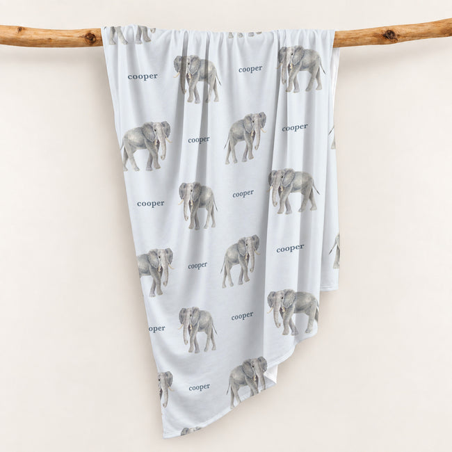 Little Elephant swaddle blanket with gray watercolor elephants and the name Cooper on pale blue, draped over a wooden branch.