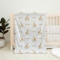 A baby blanket with a repeating pattern of brown giraffes and the name 'emerson' in brown text, draped over a light wood crib in a nursery setting.